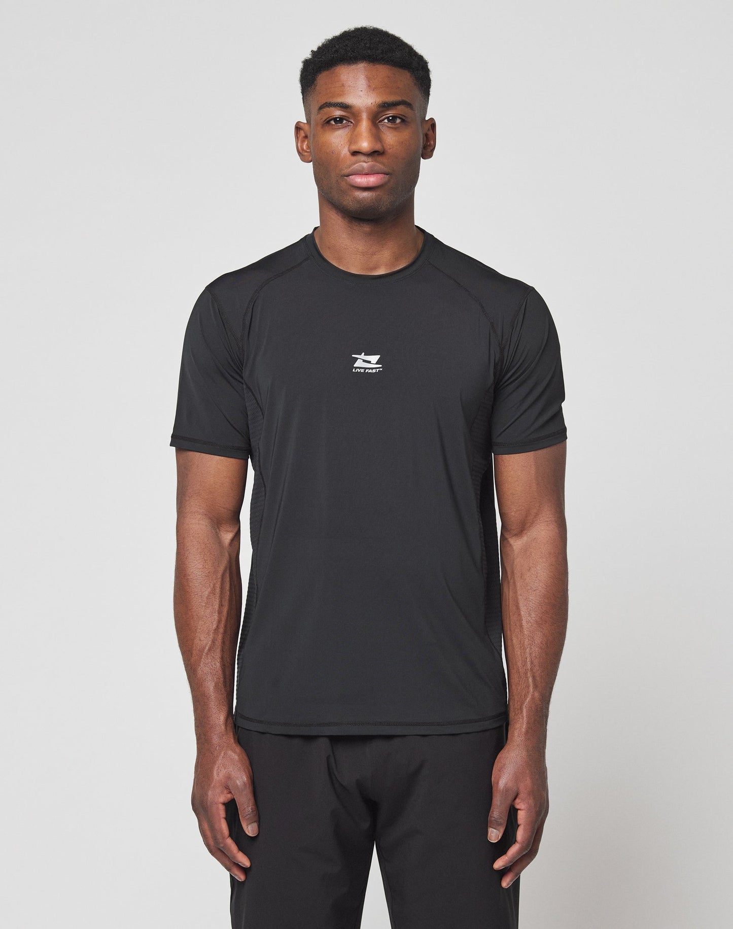 A man faces forward against a white background, wearing the LIVE FAST DIE YOUNG Live Fast Performance Tee in black, featuring a slim fit, short sleeves, small white chest logo, and comfortable Nylon Spandex material with matching black pants. | model