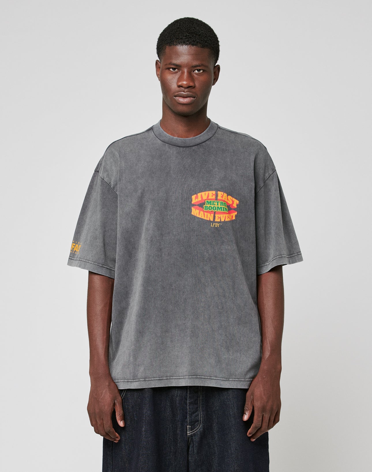A young man with short hair and a neutral expression models the LIVE FAST DIE YOUNG LFDY Metro Boomin Tee, an oversize faded gray 100% cotton shirt with yellow and red sleeve print, paired with wide dark pants against a plain background.