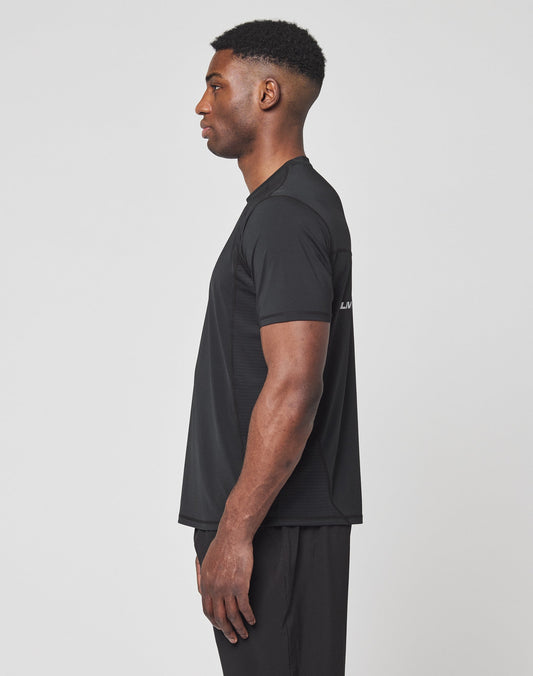 A man stands in profile facing left, wearing the LIVE FAST DIE YOUNG Live Fast Performance Tee in black and black pants against a plain white background.