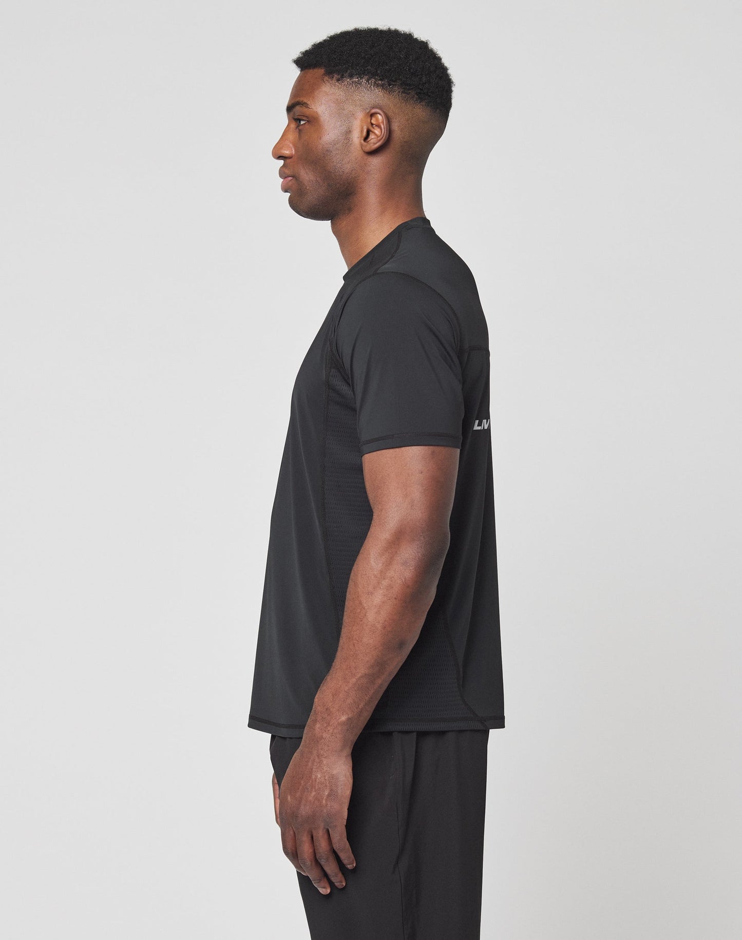 A man stands in profile facing left, wearing the LIVE FAST DIE YOUNG Live Fast Performance Tee in black and black pants against a plain white background.