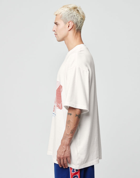 A man with short blond hair and tattoos on his left arm stands in profile wearing the LIVE FAST DIE YOUNG LFDY Juveniles Tee, an oversize vintage white 100% Baumwolle shirt with a red and blue graphic, paired with blue shorts, against a light gray background.