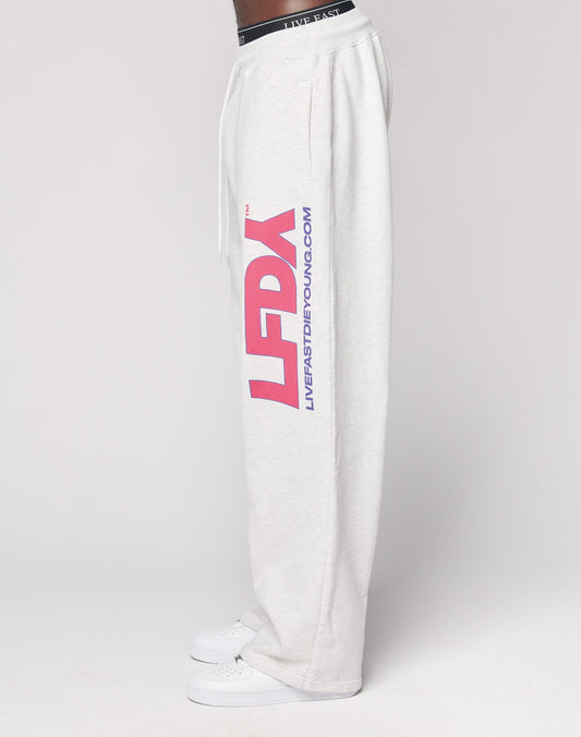 A person wears the LFDY Racing Wide Jogger by LIVE FAST DIE YOUNG—light gray sweatpants with a wide elastic waistband, bold pink LFDY lettering, blue LIVEFASTDIEYOUNG.COM on the side, styled with white sneakers, side view on a plain background.