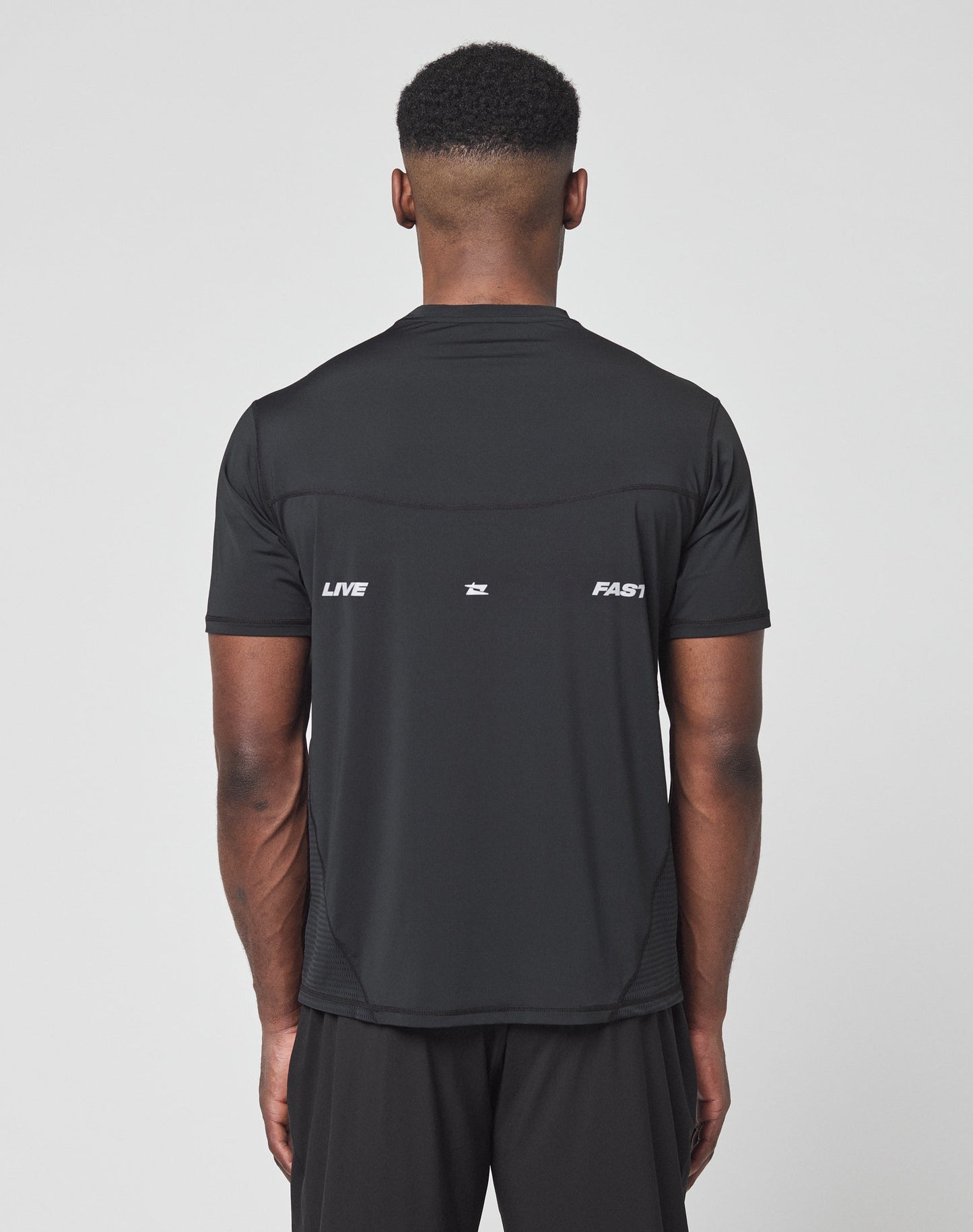 A man faces away wearing the LIVE FAST DIE YOUNG Live Fast Performance Tee, a slim-fit black nylon spandex shirt with LIVE and FAST in white on the back plus a reflective front and back logo print.