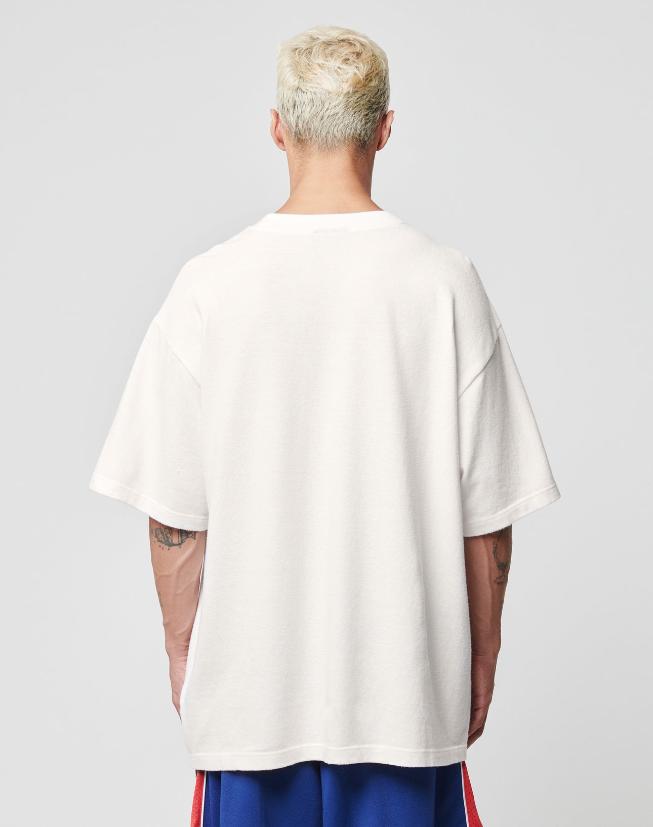 A person with short, bleached blond hair is shown from the back, wearing an oversized white LFDY Juveniles Tee by LIVE FAST DIE YOUNG and blue athletic shorts with red stripes. The background is plain light gray.