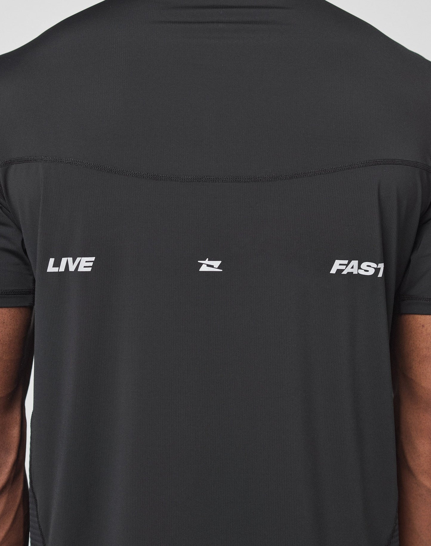 Back view of a person wearing the LIVE FAST DIE YOUNG Live Fast Performance Tee—a slim fit black Nylon Spandex shirt with LIVE on the left, FAST on the right, central logo, and reflective front/back prints.