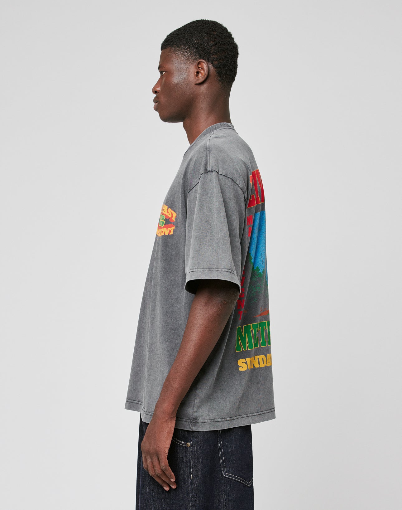 A young man in profile wears the LIVE FAST DIE YOUNG LFDY Metro Boomin Tee—an oversize fit, 100% Baumwolle faded gray shirt with colorful prints on the back and sleeve—paired with dark jeans against a plain light background.