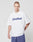 A man with short hair wears a white LIVE FAST DIE YOUNG Basic Tee (100% Baumwolle, Oversize Fit, style code LF2404-15446) with LiveFast in bold and blue athletic shorts, standing against a plain light gray background.
