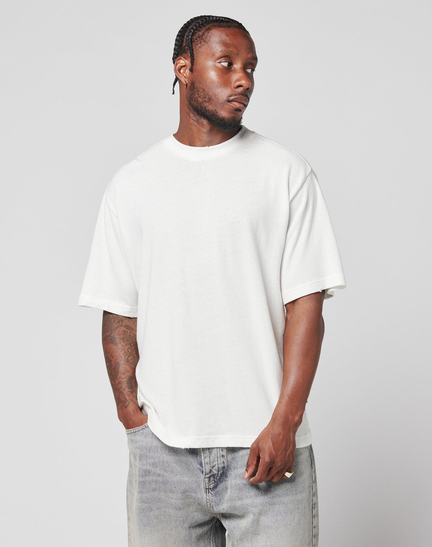 A man with braided hair wears the LIVE FAST DIE YOUNG LFDY Vintage Tee, an oversize fit 100% Baumwolle shirt, and light gray jeans. He stands against a plain light gray background, looking left with one hand in his pocket.