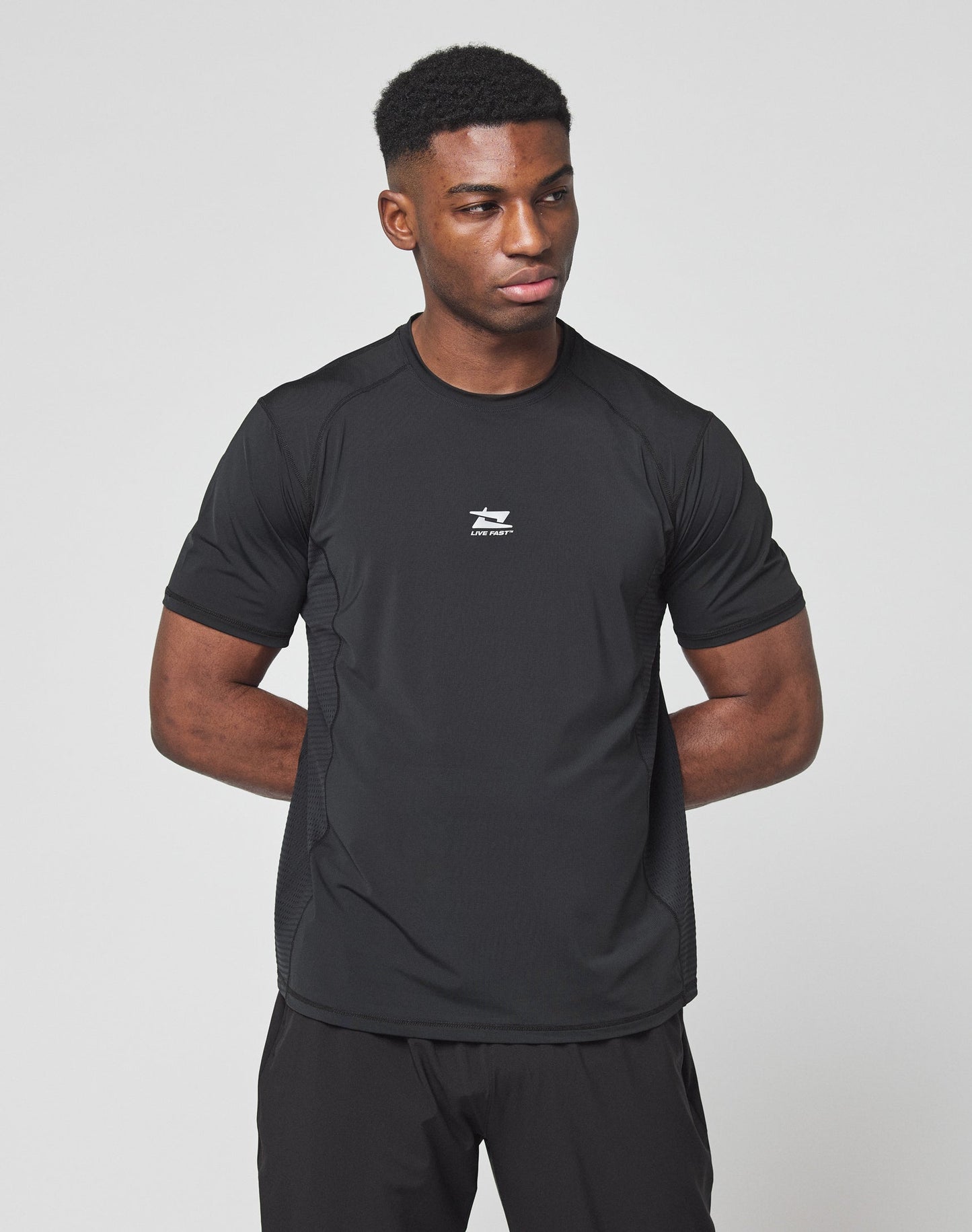 A man stands against a white background, arms behind his back, wearing the LIVE FAST DIE YOUNG Live Fast Performance Tee—a slim fit black athletic shirt with a small white chest logo, made from comfortable nylon spandex material.
