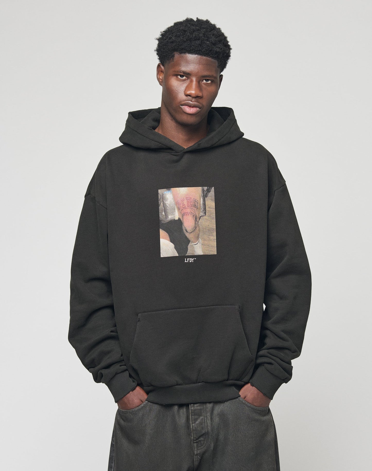 A young man with short curly hair wears the LFDY Sharkhunter Hooded by LIVE FAST DIE YOUNG, featuring front and back prints, posing with his hands in his pockets against a plain light background.