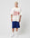 A person with short blonde hair stands against a plain white background, wearing an oversize fit LIVE FAST DIE YOUNG LFDY Juveniles Tee in vintage white with red text, blue athletic shorts with side stripes, white crew socks, and white sneakers.