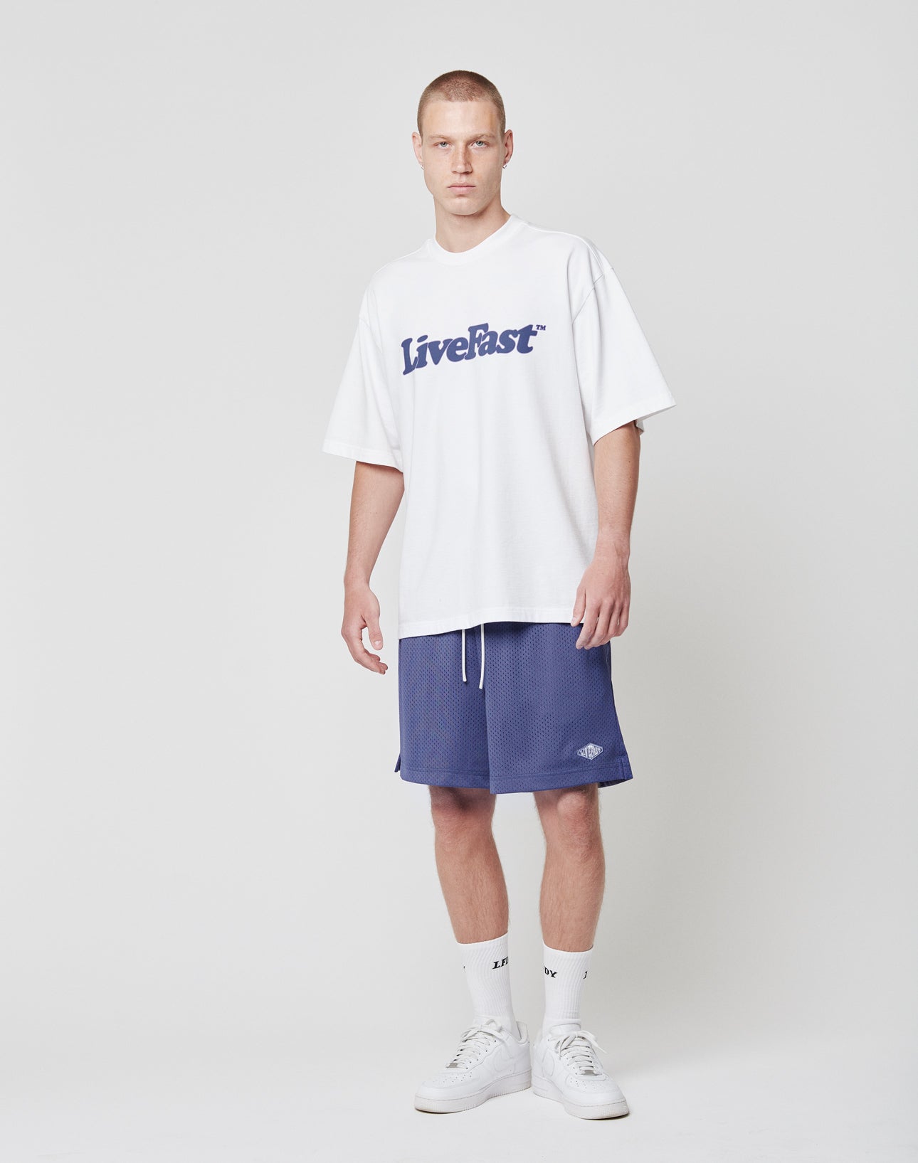 A man stands against a plain white background wearing a white oversized LIVE FAST DIE YOUNG T-shirt with logo patch, blue Basic Mesh Shorts, white crew socks, and white sneakers. He faces the camera with a neutral expression.