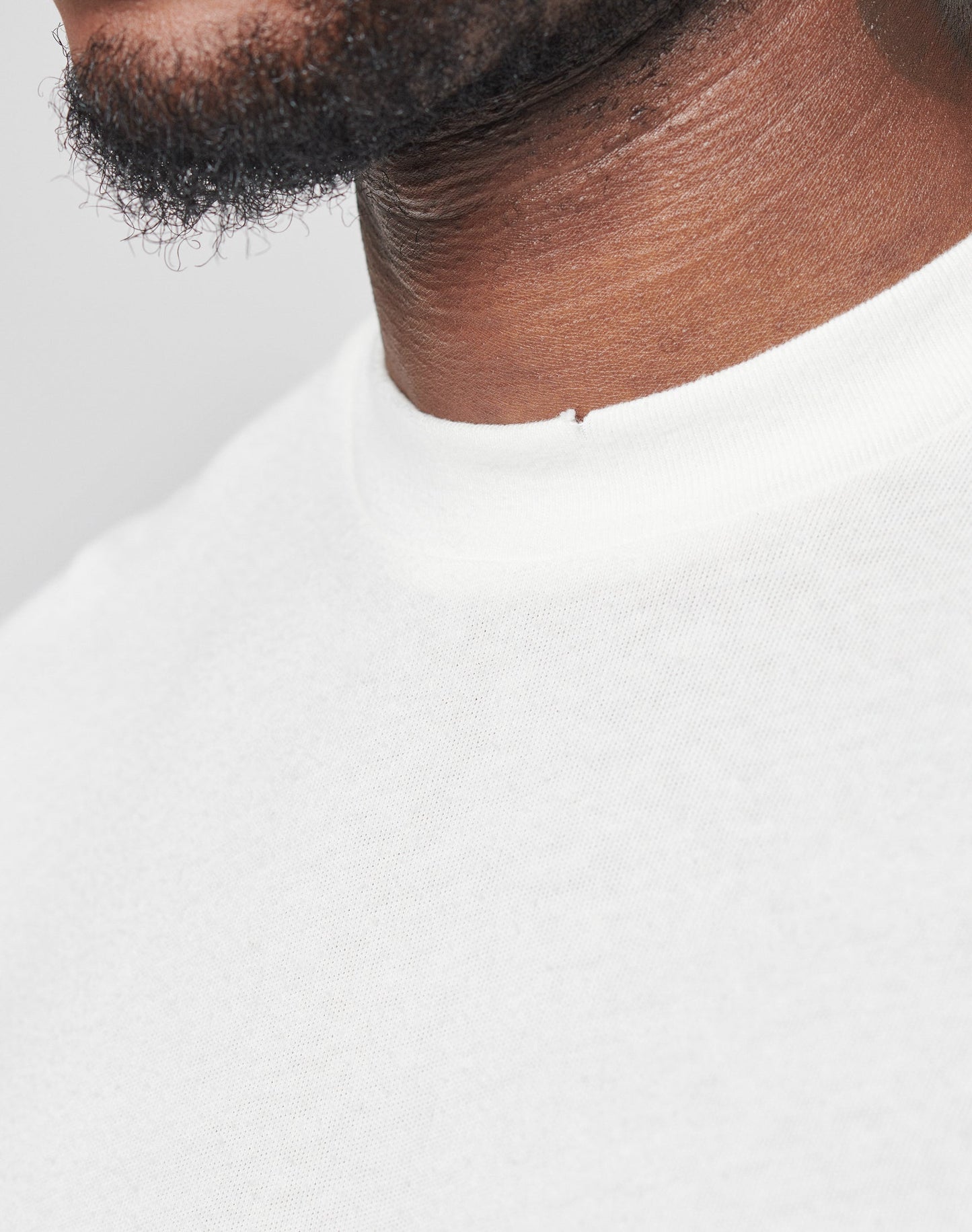 Close-up of the lower face and neck of a person with a short beard and dark skin, wearing the LFDY Vintage Tee by LIVE FAST DIE YOUNG—an oversize fit plain white crew-neck shirt made from 100% cotton—against a light background.