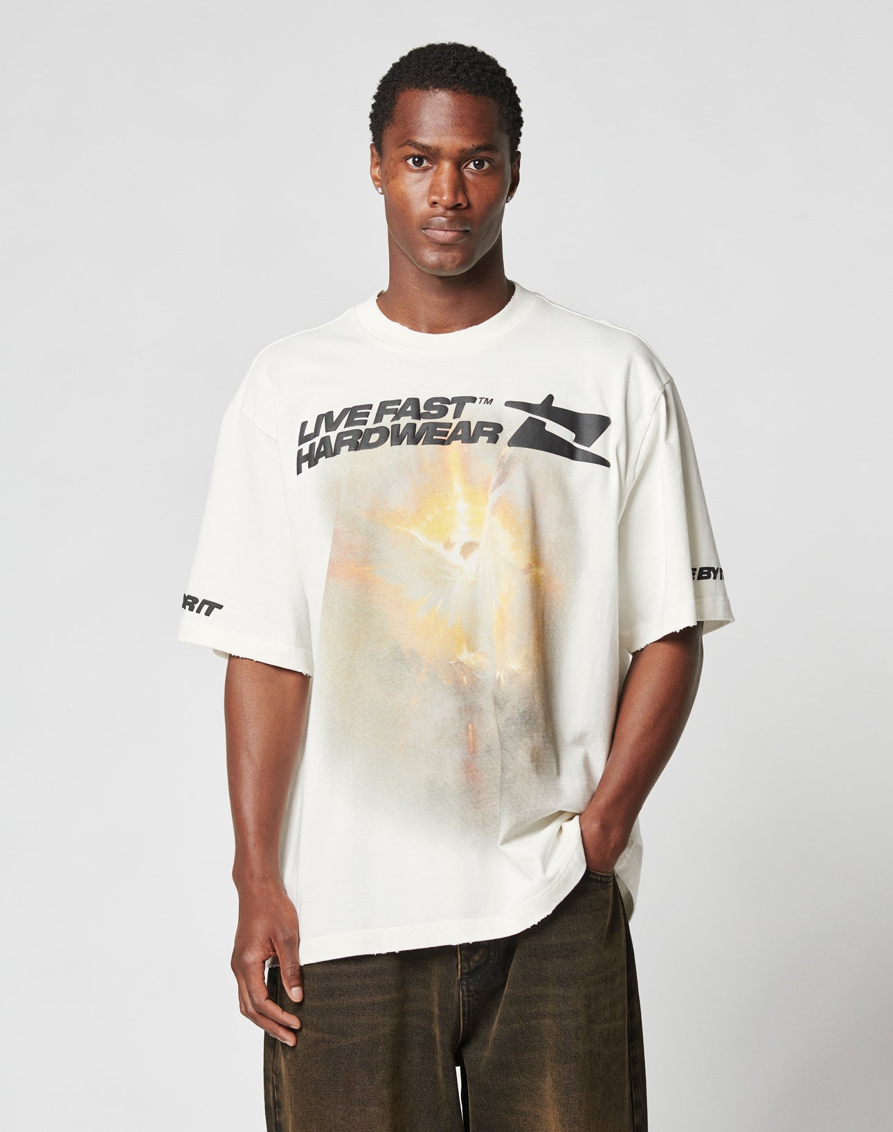 A man with short hair wears the LIVE FAST DIE YOUNG LFDY Angel Tee, an oversize-fit white T-shirt made from 100% Baumwolle with black LIVE FAST HARDWEAR text and graphic prints on the front and sleeves. He pairs it with dark pants against a plain light background.