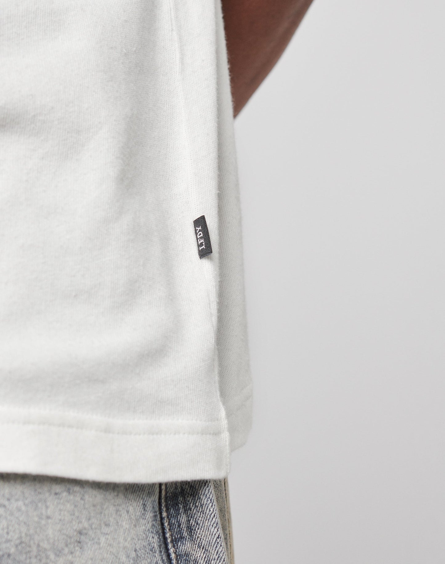 Close-up of a person wearing the LFDY Vintage Tee by LIVE FAST DIE YOUNG in white, featuring an oversize fit, 100% cotton fabric, and a small black ROT tag near the hem. Styled with light blue jeans against a plain light background.