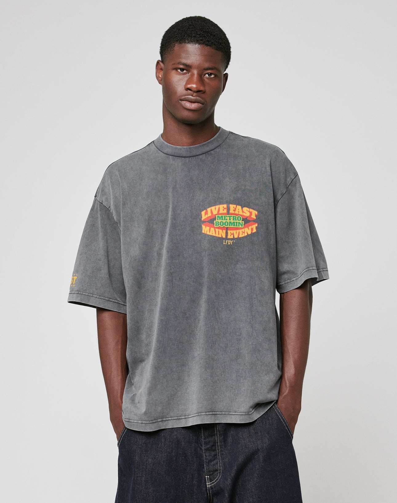 A man with a neutral expression stands against a plain background in the LIVE FAST DIE YOUNG LFDY Metro Boomin Tee—an oversize fit, 100% Baumwolle faded gray shirt with a colorful Live Fast Main Event chest graphic—and loose dark jeans. Hands in pockets.