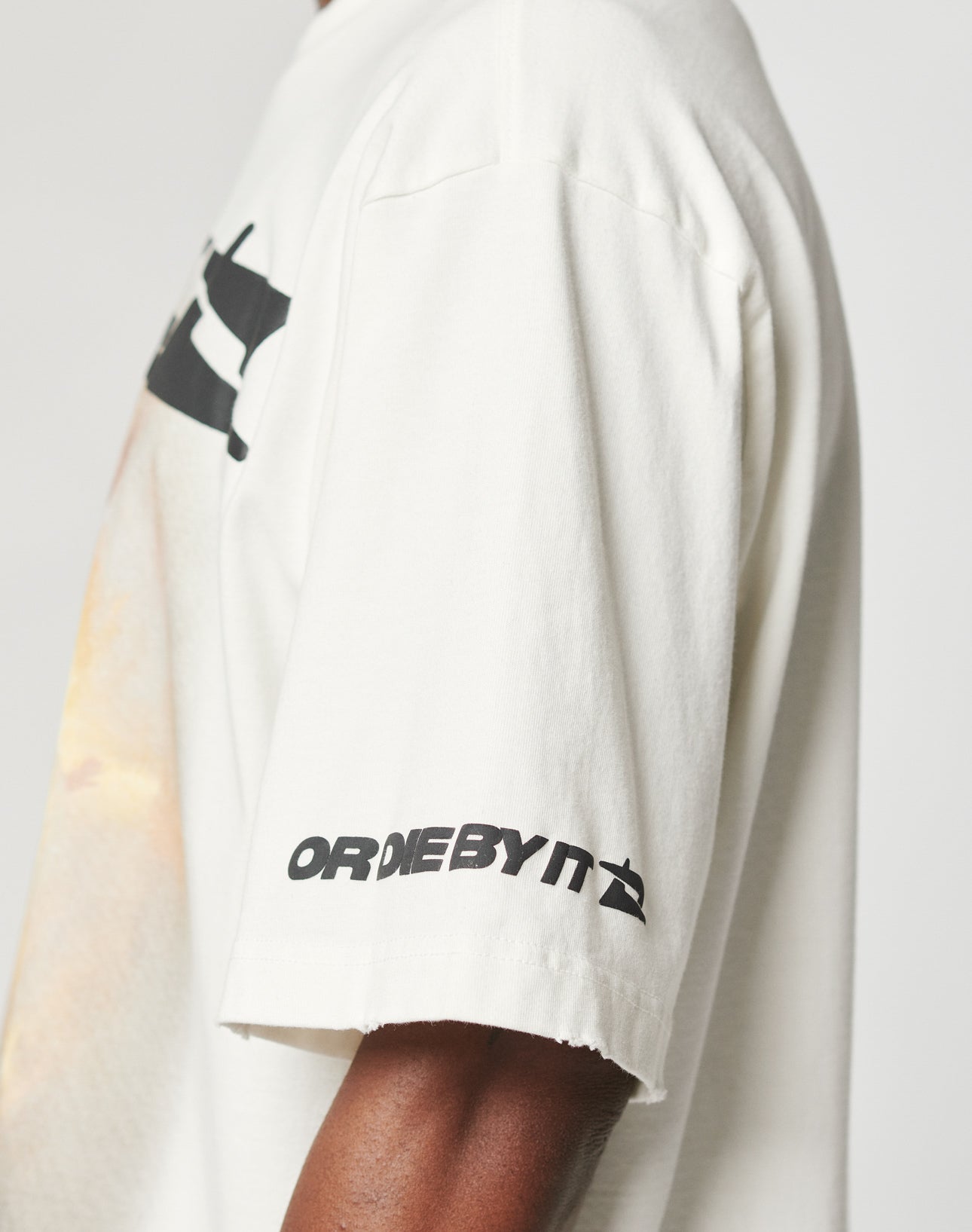 A person wears the LFDY Angel Tee by LIVE FAST DIE YOUNG, an oversize off-white 100% Baumwolle T-shirt with black ORDERBYIT text and prints on the front and sleeves. The image shows a side view of their upper body against a plain background.