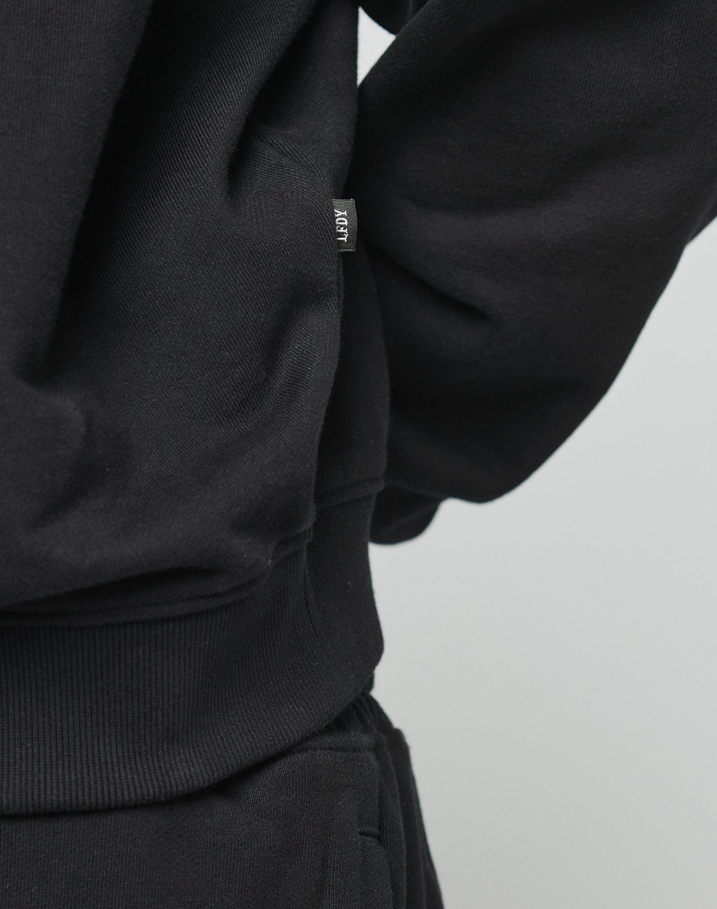 Close-up of a person wearing the DY Sweater by LIVE FAST DIE YOUNG in black, featuring an oversize fit and raglan sleeves, paired with black pants. A small white ARHT tag is visible on the side seam. The background is plain and light-colored.