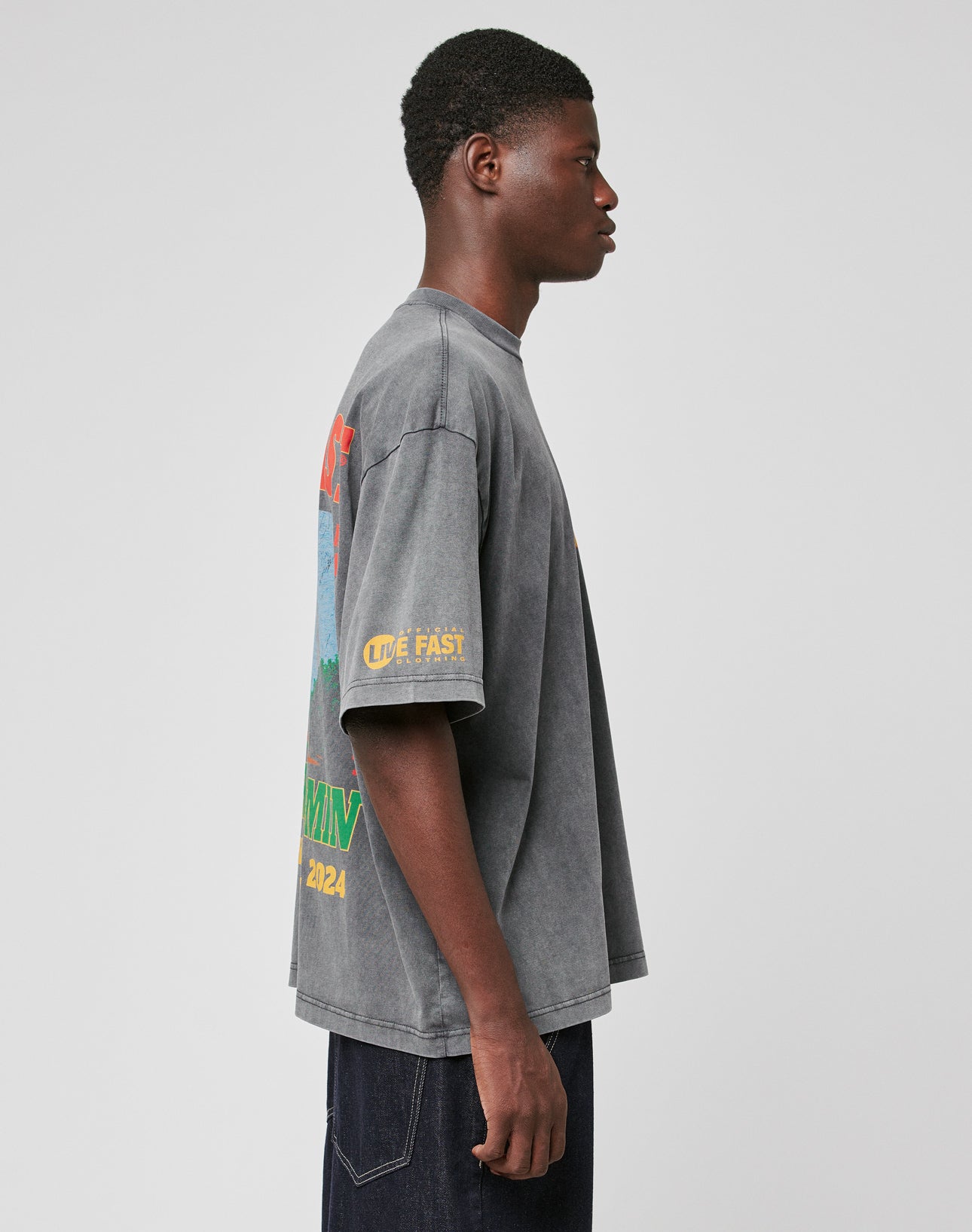 A young man stands sideways in a LIVE FAST DIE YOUNG LFDY Metro Boomin Tee, an oversize fit faded gray graphic shirt with colorful text and sleeve print, made of 100% Baumwolle, paired with dark jeans. The background is plain light gray.