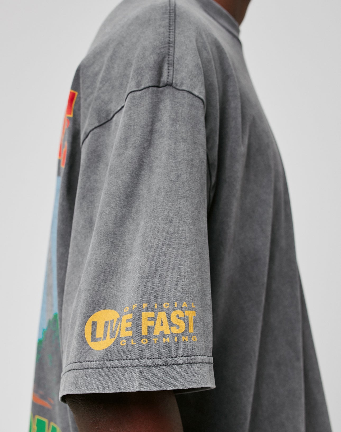 A person wears the LIVE FAST DIE YOUNG LFDY Metro Boomin Tee in oversized faded grey 100% cotton with a yellow Official Live Fast Clothing print on the sleeve, shown from the side against a plain background.