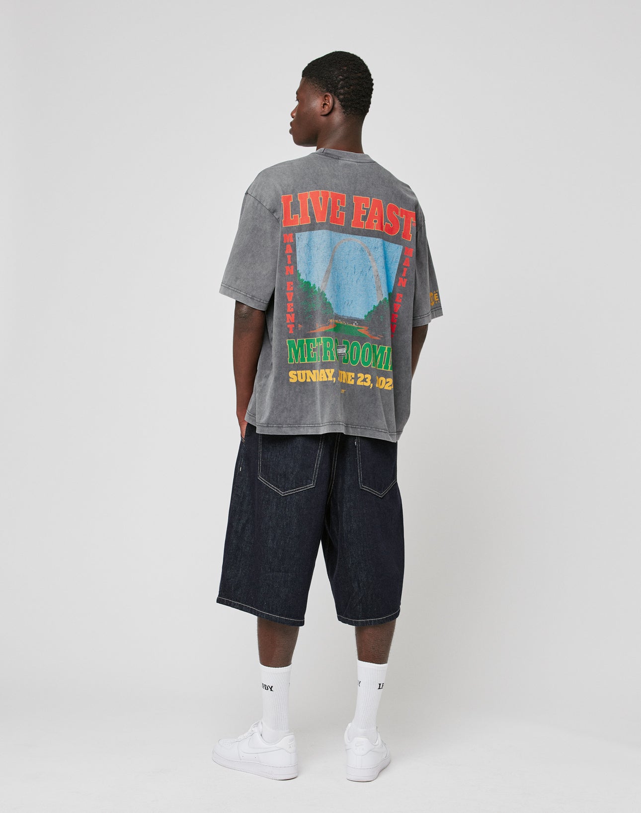 A man stands facing away, wearing the LFDY Metro Boomin Tee by LIVE FAST DIE YOUNG—a gray oversize fit T-shirt with colorful back graphics—paired with dark baggy denim shorts, white socks, and sneakers against a plain light background.