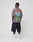 A man stands facing away, wearing the LFDY Metro Boomin Tee by LIVE FAST DIE YOUNG—a gray oversize fit T-shirt with colorful back graphics—paired with dark baggy denim shorts, white socks, and sneakers against a plain light background.