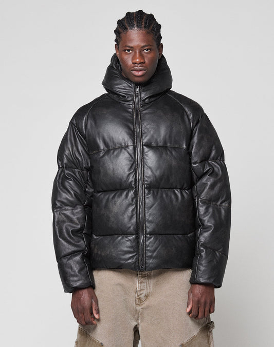 A person stands against a plain background wearing the LIVE FAST DIE YOUNG Live Fast Leather Puffer Jacket—black, oversized with a hood and embossed logo—paired with light brown, loose-fitting pants and neatly braided hair. | model