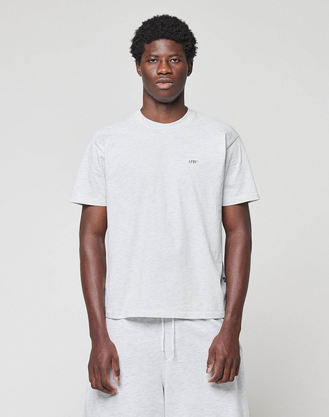 A man stands against a plain light background, wearing a light gray Basic Chest Tee by LIVE FAST DIE YOUNG and matching gray shorts. This regular-fit T-shirt is made from 100% Baumwolle and features small text on the left chest. | model