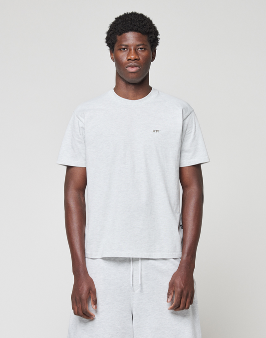 A man stands against a plain light background, wearing a light gray Basic Chest Tee by LIVE FAST DIE YOUNG and matching gray shorts. This regular-fit T-shirt is made from 100% Baumwolle and features small text on the left chest. | model