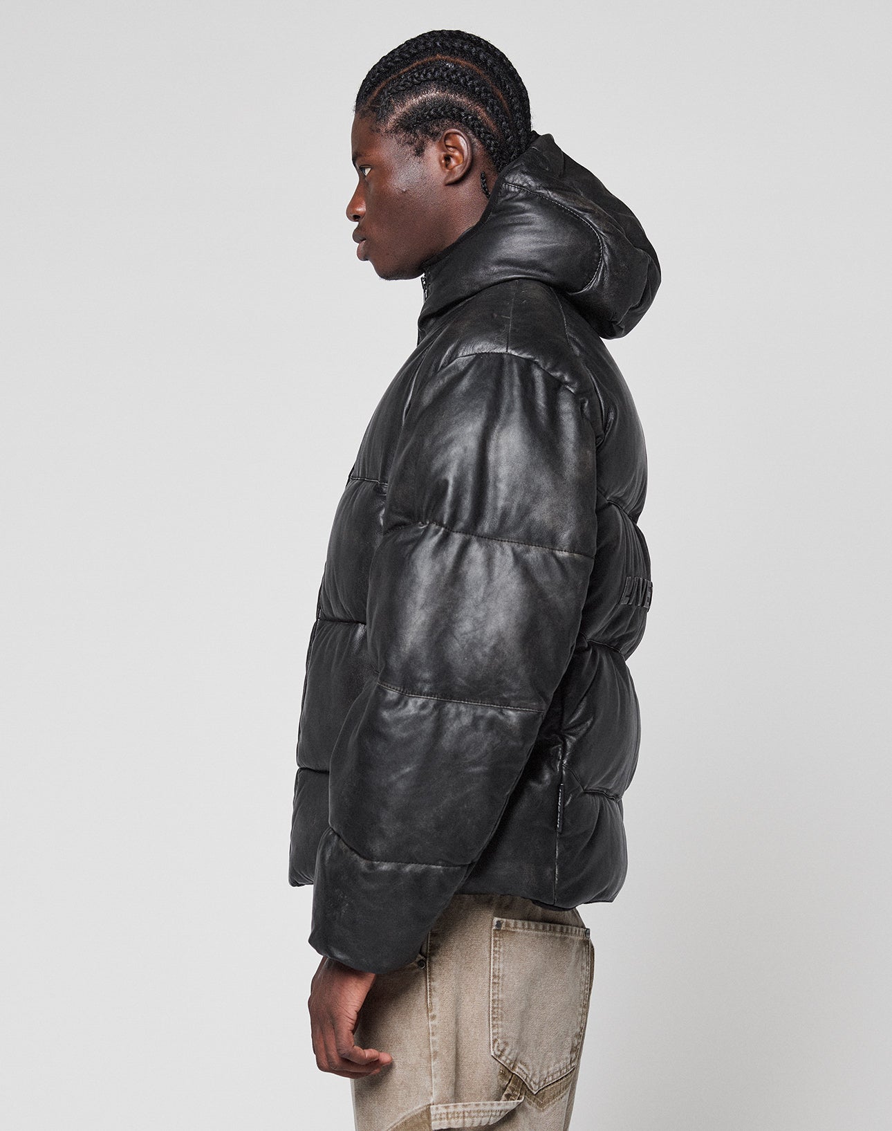 A person with braided hair stands in profile against a light gray background, wearing the LIVE FAST DIE YOUNG Live Fast Leather Puffer Jacket—black, shiny, embossed logo with hood—and light brown pants.