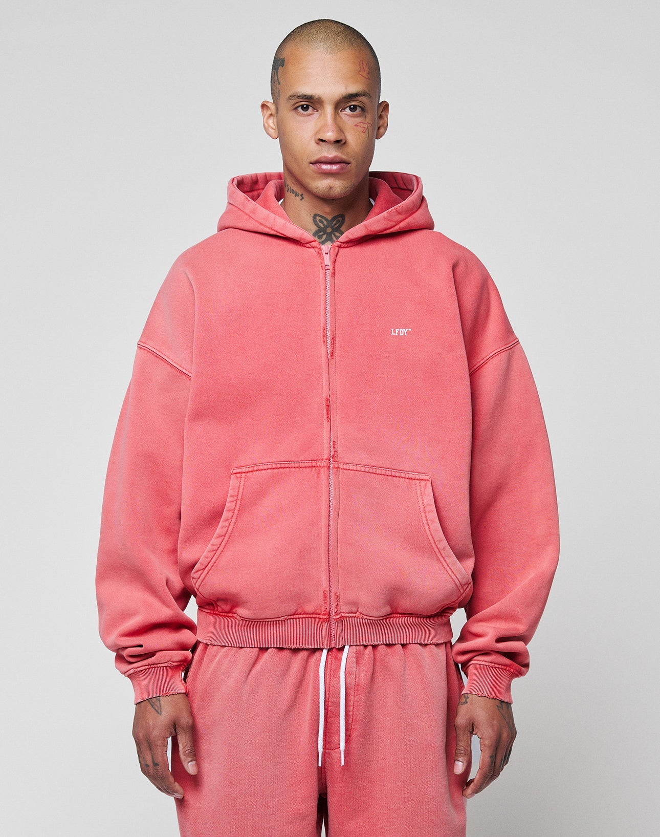 A person with a shaved head and neck tattoos wears the LIVE FAST DIE YOUNG Basic Hooded Zip and matching sweatpants in pink with a subtle fade effect, standing against a plain light gray background. | model