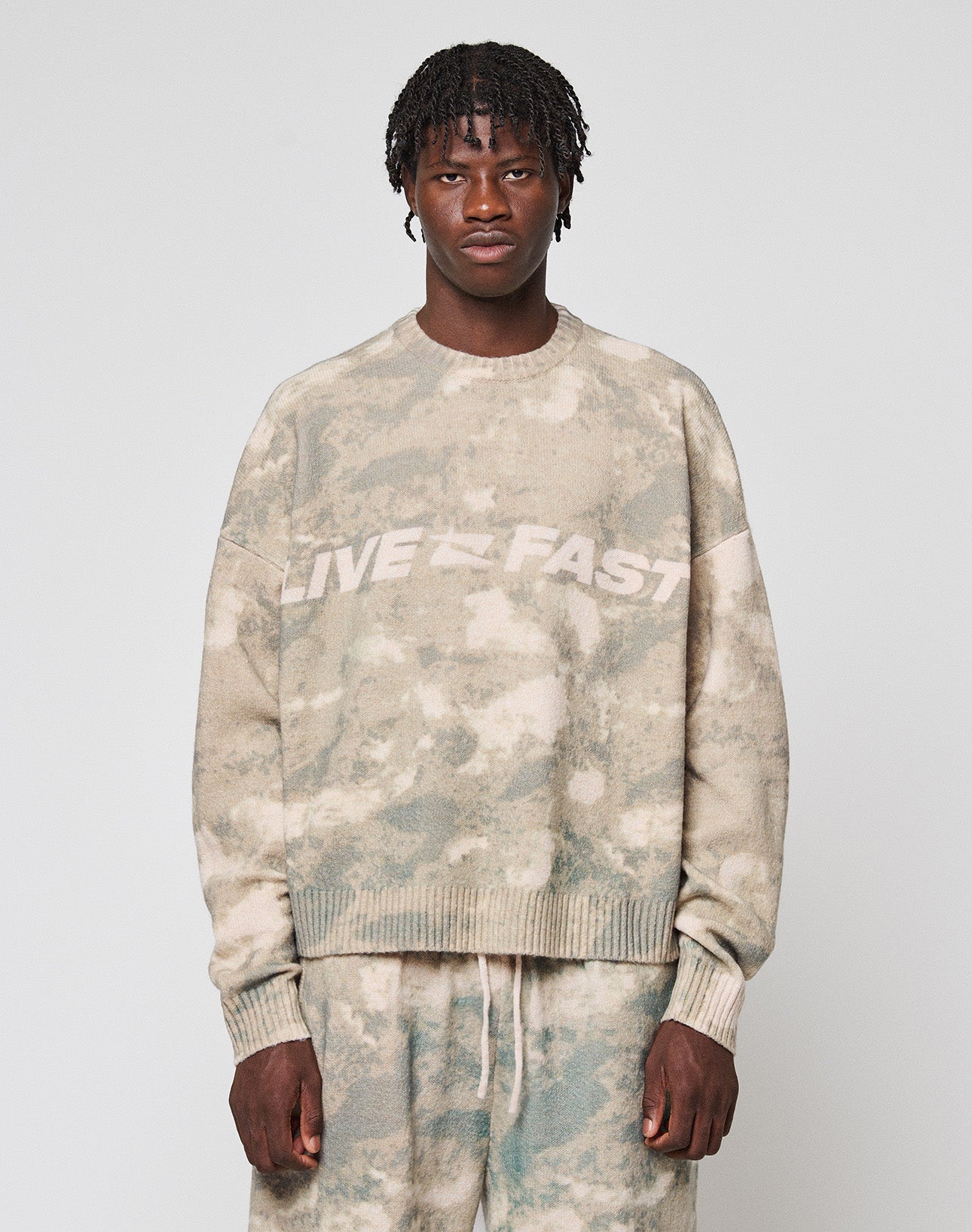 A person faces forward wearing the LIVE FAST DIE YOUNG Live Fast Boucle Sweater in an oversized beige and green tie-dye design with "LIVE FAST" on the chest, paired with matching soft polyester pants against a plain light gray background. | model