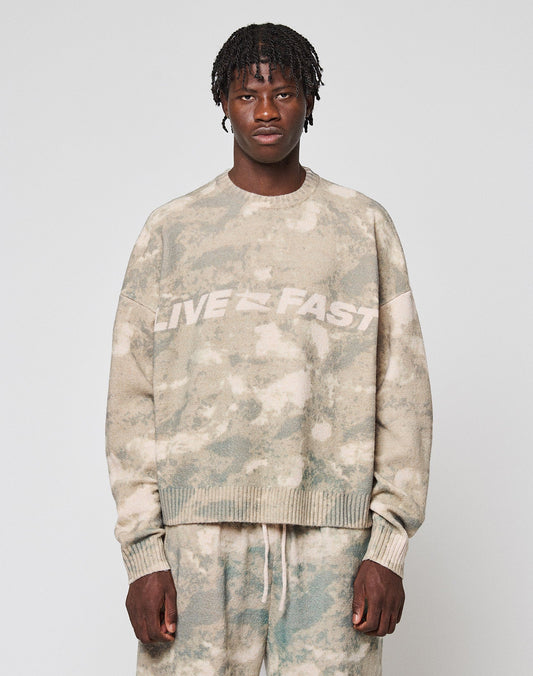 A person faces forward wearing the LIVE FAST DIE YOUNG Live Fast Boucle Sweater in an oversized beige and green tie-dye design with "LIVE FAST" on the chest, paired with matching soft polyester pants against a plain light gray background. | model