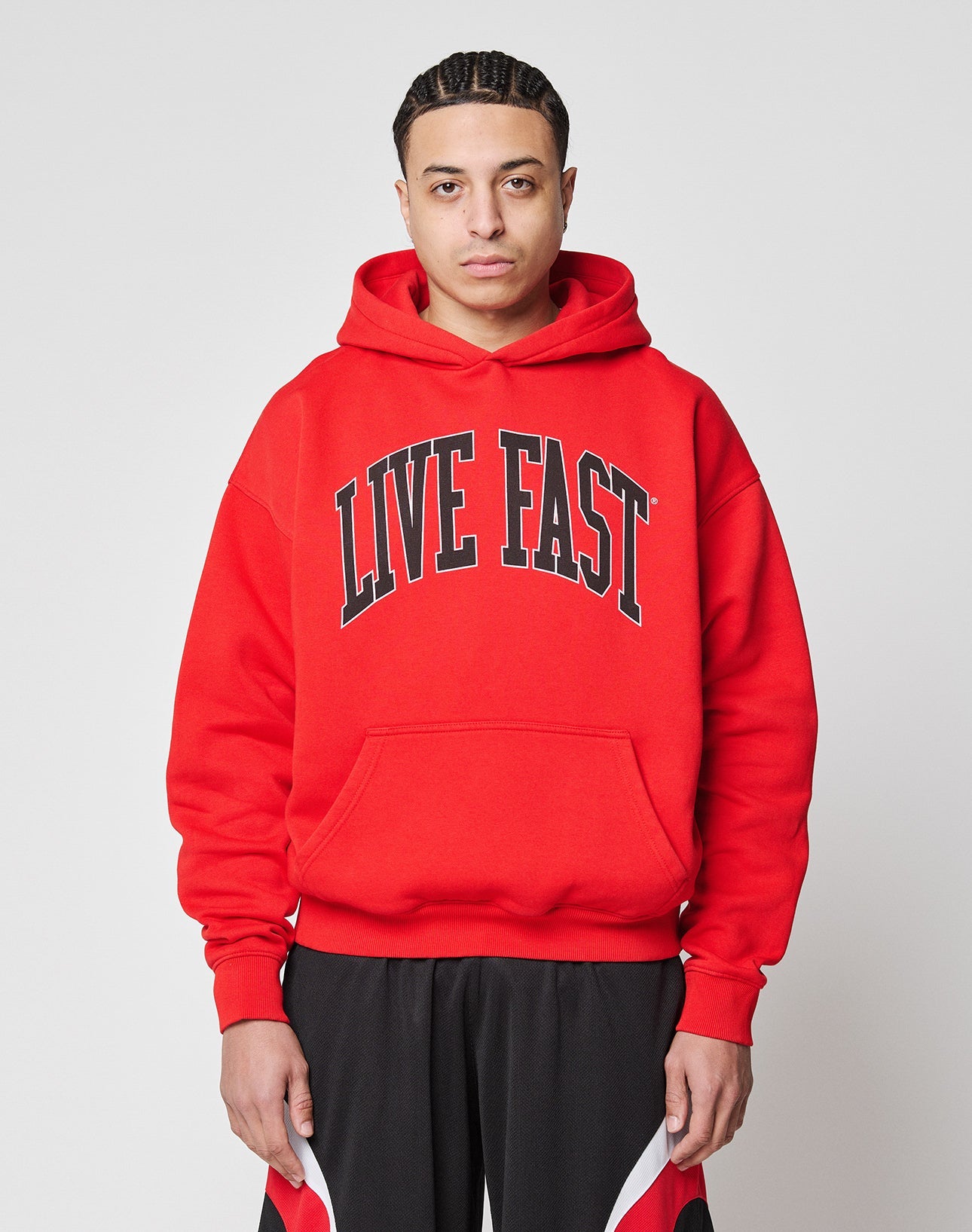 A person faces the camera in an oversized bright red Live Fast College Hooded by LIVE FAST DIE YOUNG, featuring bold black LIVE FAST text and black/red athletic shorts, made from a comfortable polyester blend, against a plain white background.