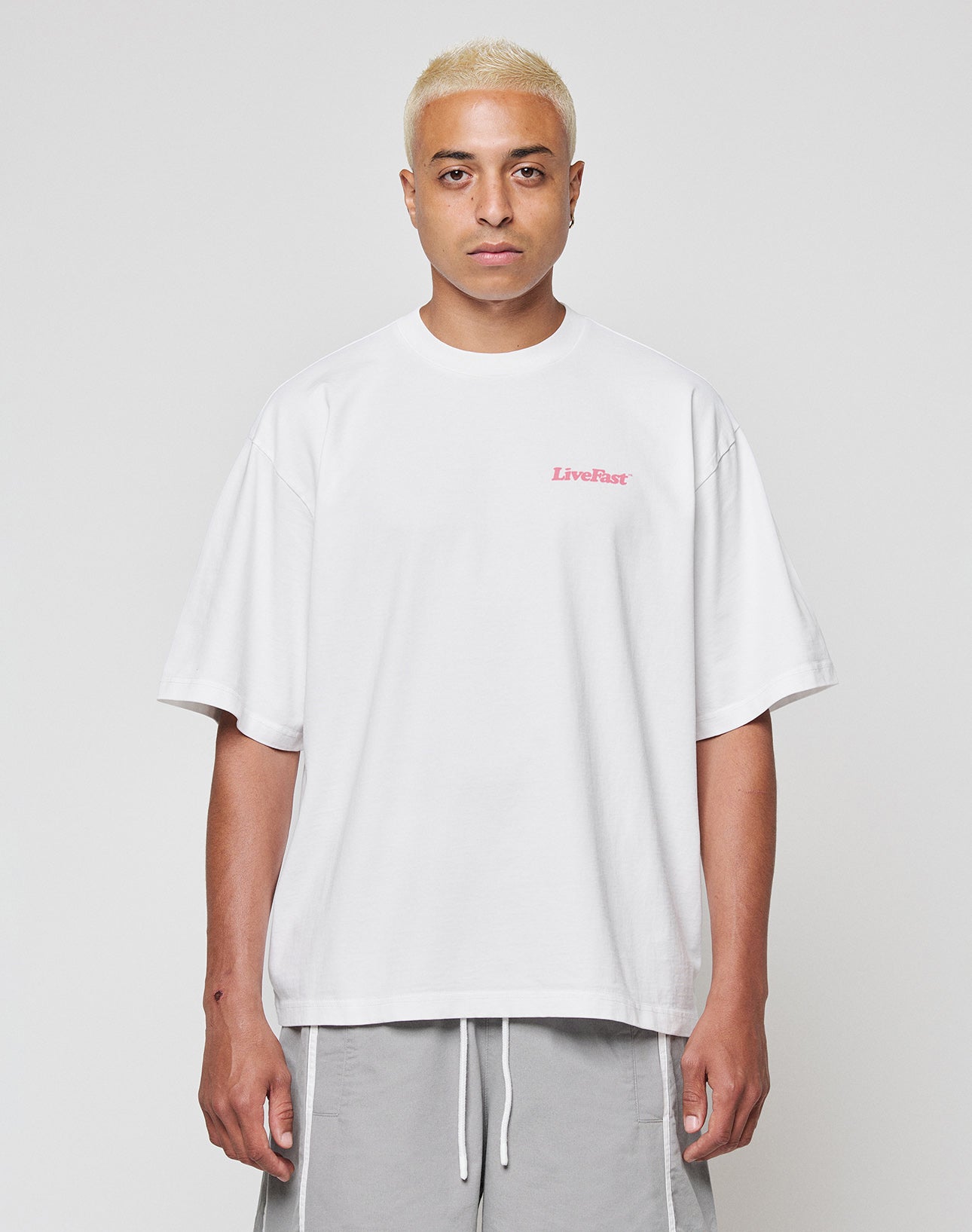 A person with short bleached blonde hair wears the LIVE FAST DIE YOUNG Basic 280 Tee, an oversize white 100% Baumwolle shirt with small pink LiveFast text on the chest, and light gray shorts, standing against a plain white background.