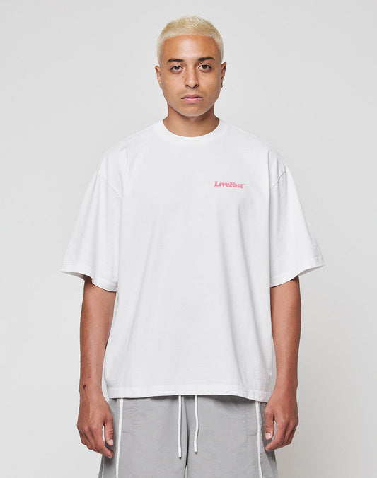 A person with short bleached blonde hair wears the LIVE FAST DIE YOUNG Basic 280 Tee, an oversize white 100% Baumwolle shirt with small pink LiveFast text on the chest, and light gray shorts, standing against a plain white background.