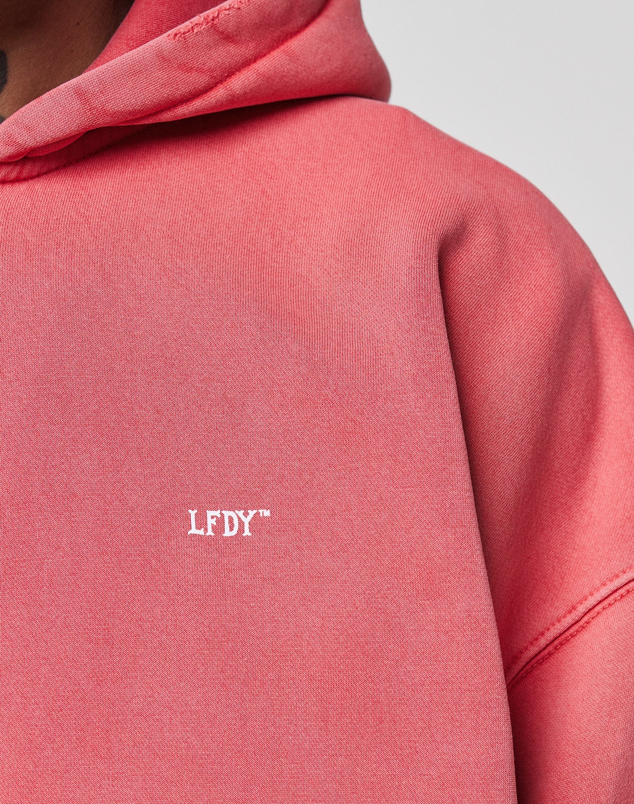 Close-up of a person wearing the LIVE FAST DIE YOUNG Basic Hooded Zip in pink, featuring a small white LFDY™ logo print on the front and showcasing the fabric’s subtle fade effect. Only part of the hoodie and their neck and chin are visible.