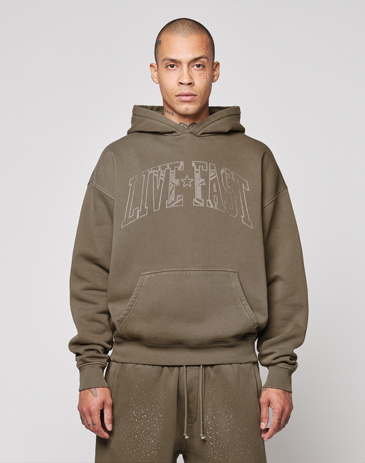 A person faces the camera against a plain background, wearing the LIVE FAST DIE YOUNG Live Fast Rhinestone Hooded in olive green with an oversize fit and "LIVE FAST" on the front, paired with matching speckled sweatpants. | model