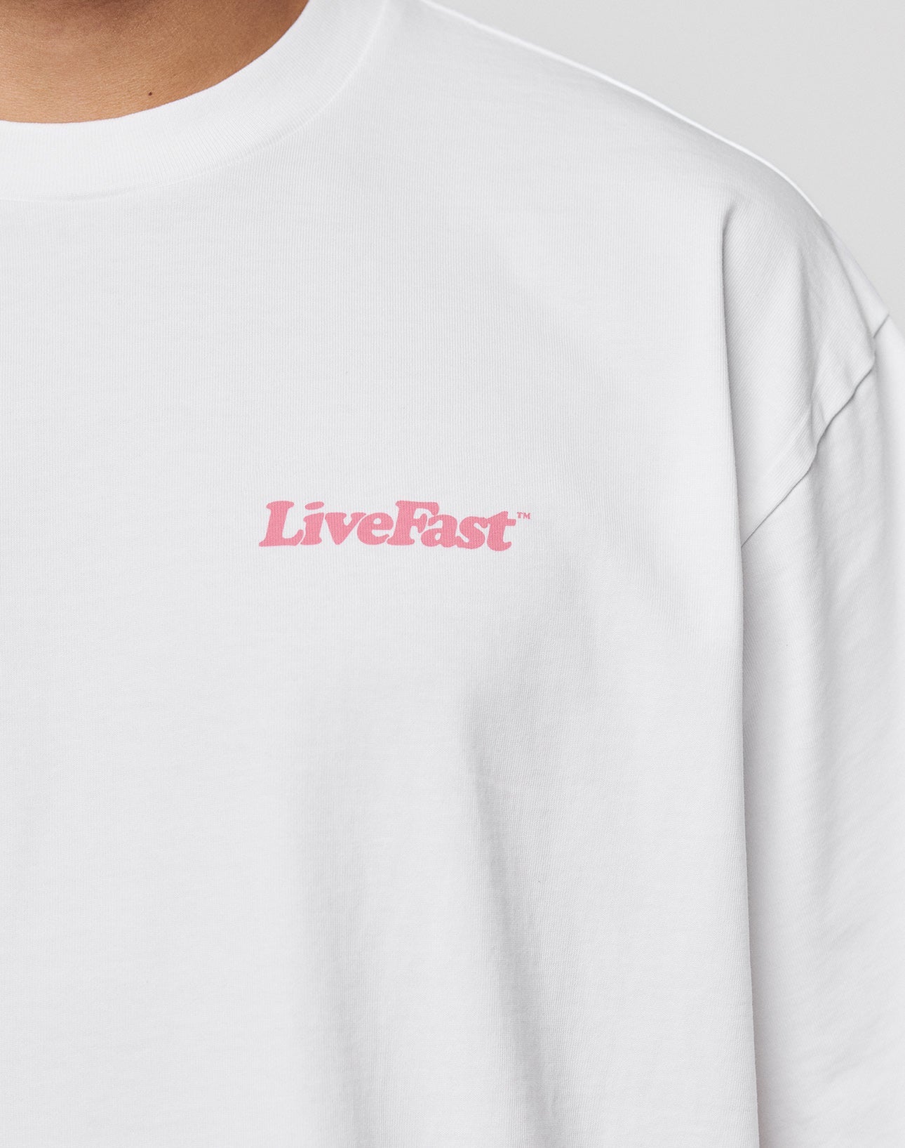 Close-up of a person wearing the LIVE FAST DIE YOUNG Basic 280 Tee, an oversize fit white t-shirt made from 100% Baumwolle, with LiveFast in pink text on the upper left chest.