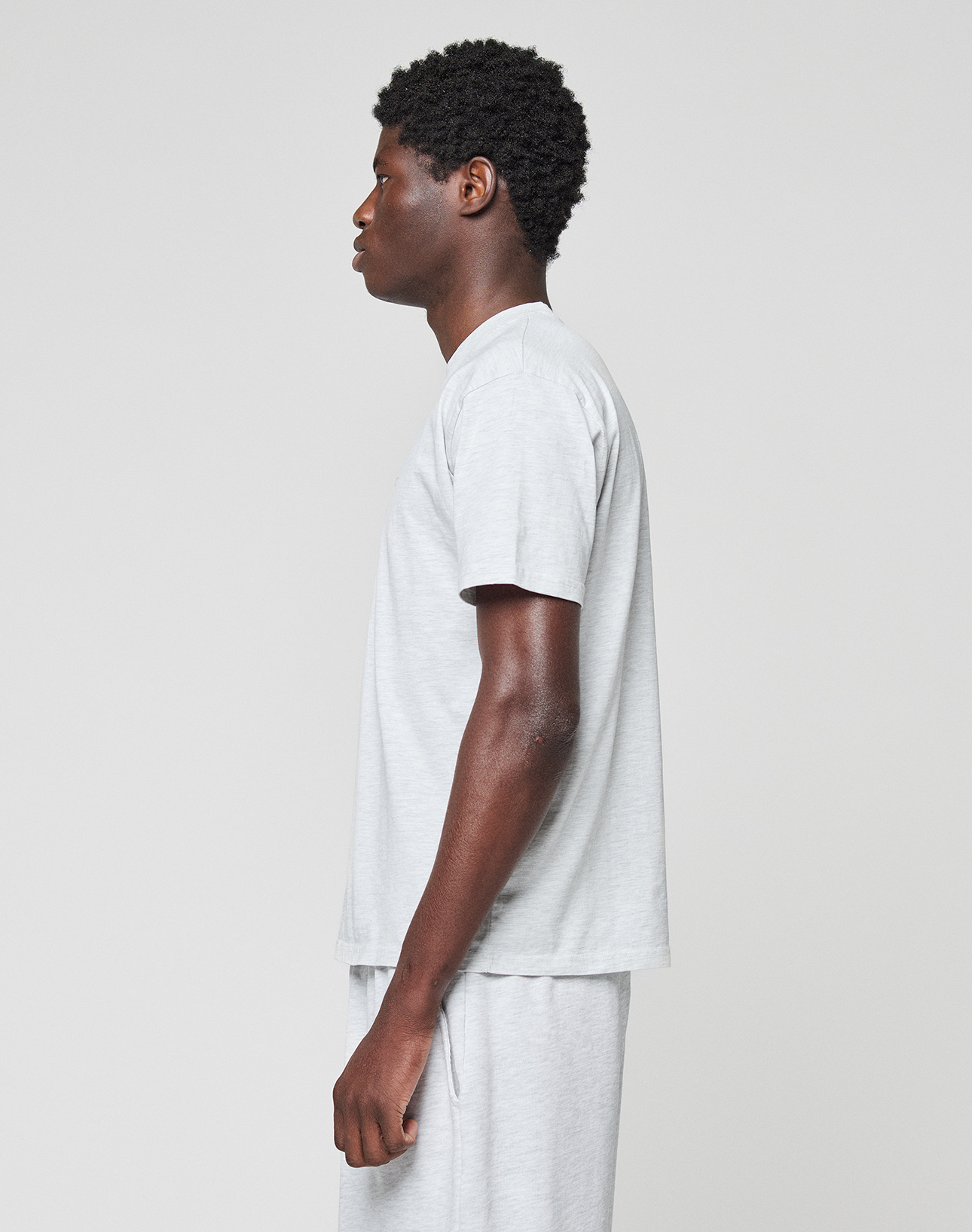 A person with short curly hair stands in profile facing left, wearing a light gray LIVE FAST DIE YOUNG Basic Chest Tee and matching shorts, both made from 100% Baumwolle, against a plain light background.