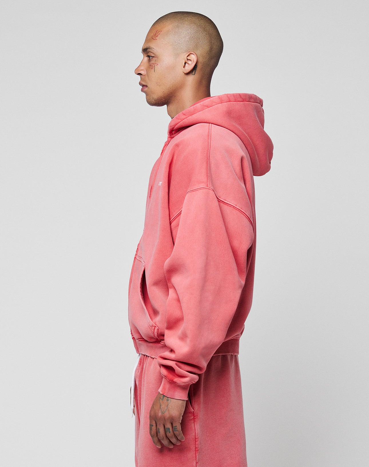 A shaved-headed person stands in profile, wearing the LIVE FAST DIE YOUNG Basic Hooded Zip in faded pink with matching sweatpants, set against a plain light gray background.