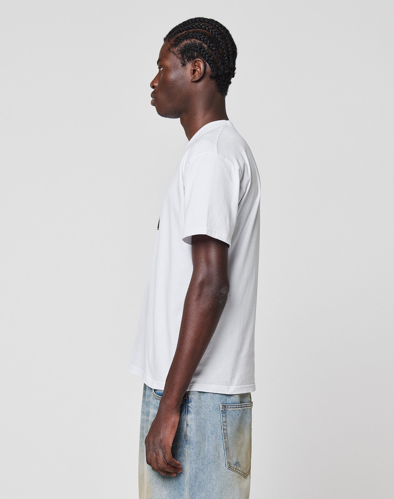 A person with braided hair stands in profile facing left, wearing a regular fit LIVE FAST DIE YOUNG LFDY Gambling Tee and light blue jeans against a plain light gray background.