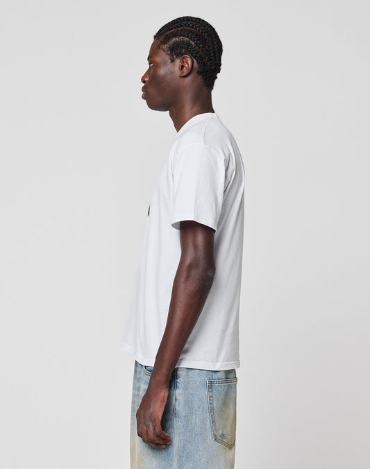 A person with braided hair stands in profile facing left, wearing a regular fit LIVE FAST DIE YOUNG LFDY Gambling Tee and light blue jeans against a plain light gray background.