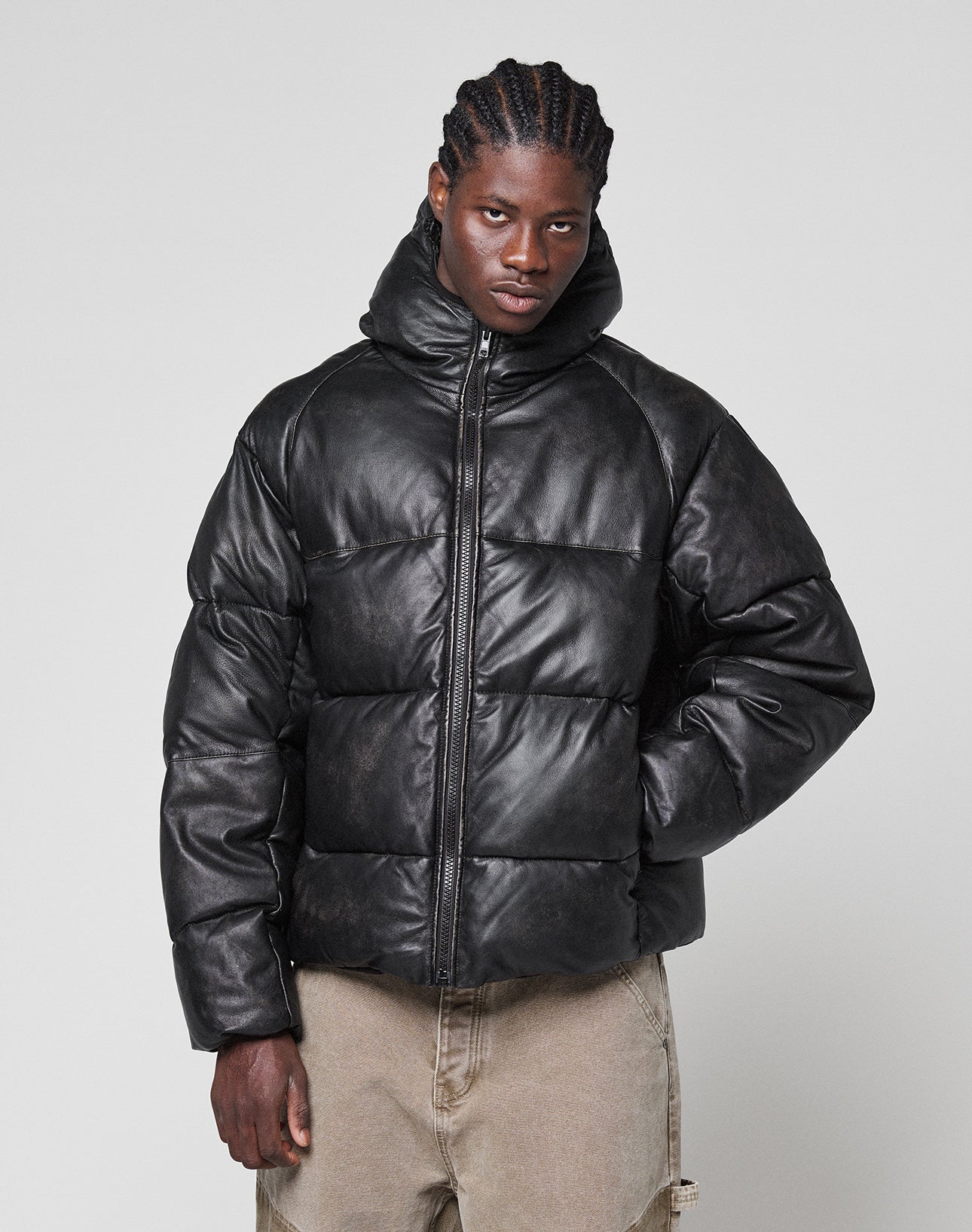 A man with braided hair wears the Live Fast Leather Puffer Jacket by LIVE FAST DIE YOUNG, featuring an oversize fit and high collar, styled with tan pants against a plain light gray background.