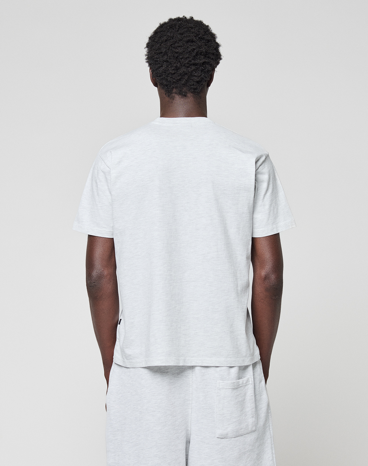 A person with short curly hair stands with their back to the camera, wearing a light gray LIVE FAST DIE YOUNG Basic Chest Tee and matching shorts against a plain light background.