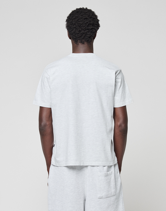 A person with short curly hair stands with their back to the camera, wearing a light gray LIVE FAST DIE YOUNG Basic Chest Tee and matching shorts against a plain light background.
