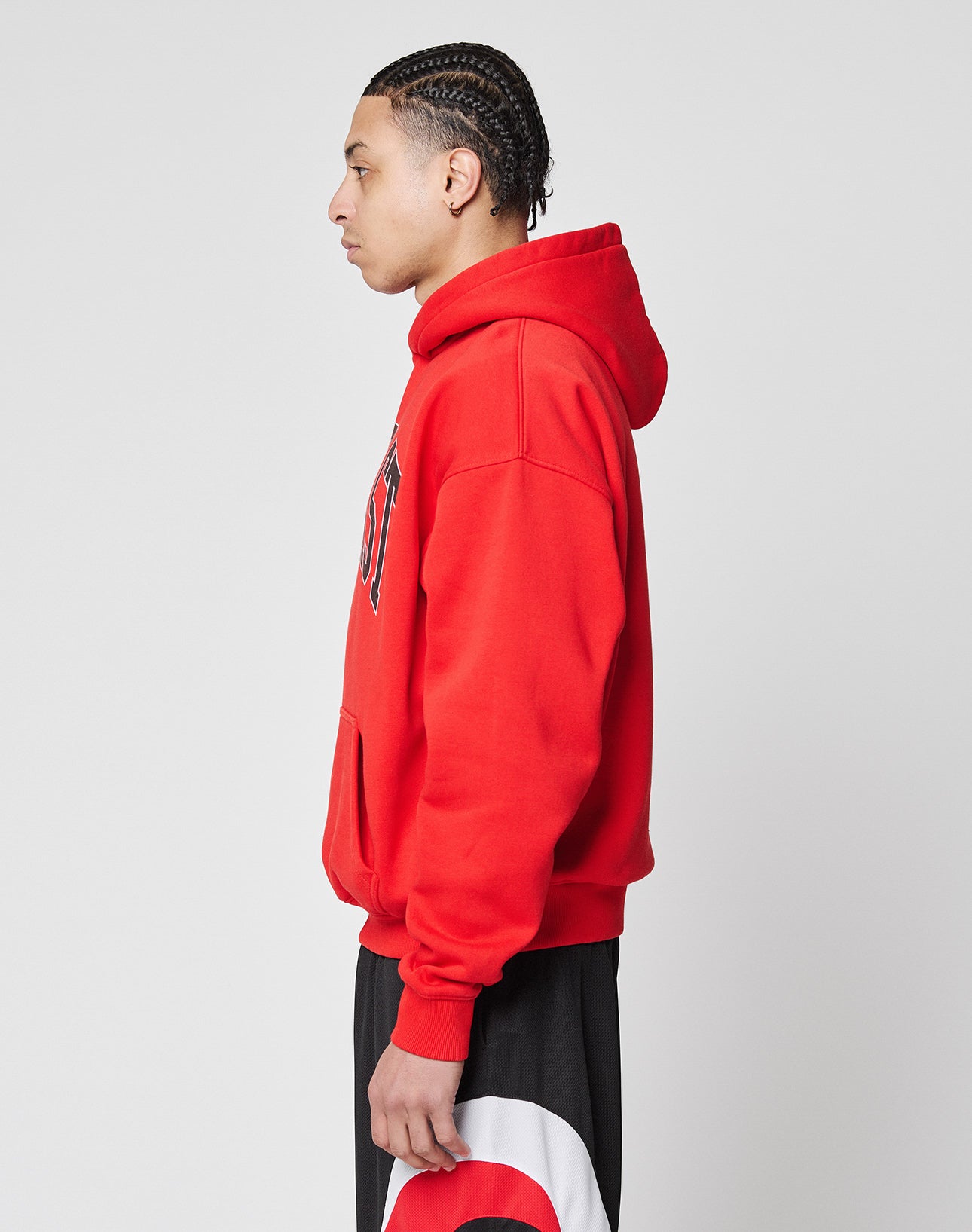 A young man with braided hair stands in profile against a white background, wearing a bright red LIVE FAST DIE YOUNG Live Fast College Hooded sweatshirt and black and white pants with a red design.