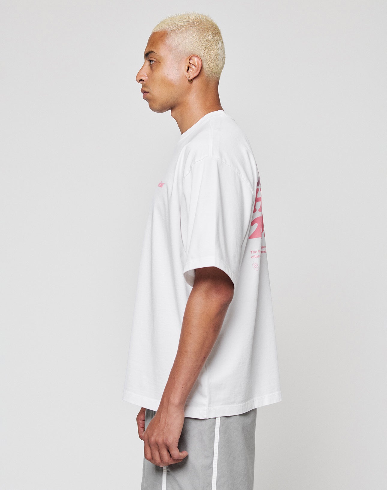 A person with short bleached blond hair stands in profile against a plain background, wearing the LIVE FAST DIE YOUNG Basic 280 Tee in white with pink front and back print, paired with light gray shorts featuring a white stripe.