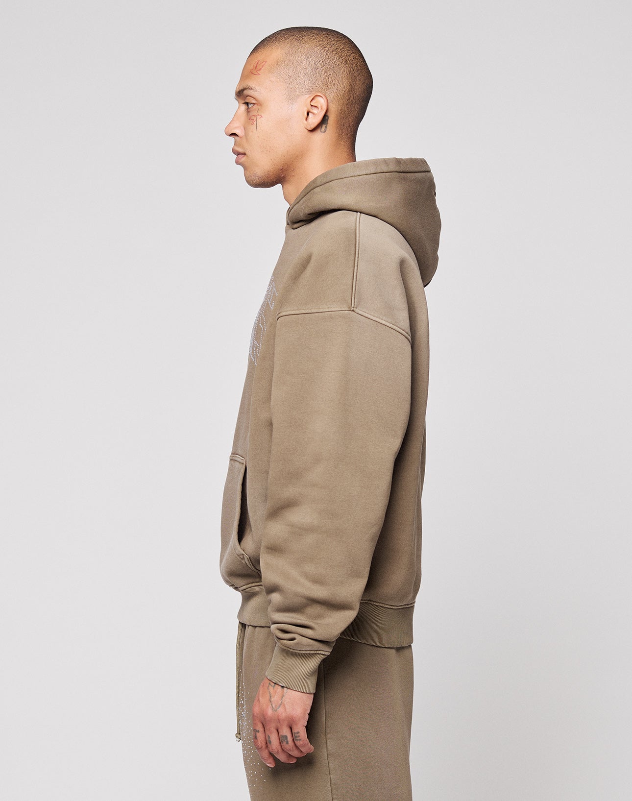 A person with short hair stands in profile wearing the LIVE FAST DIE YOUNG Live Fast Rhinestone Hooded in taupe, styled with matching oversized sweatpants against a plain light background.