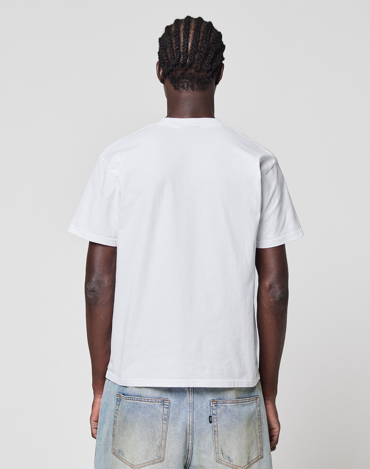A person with braided hair faces away from the camera, wearing the LIVE FAST DIE YOUNG LFDY Gambling Tee aus 100% Baumwolle and light blue jeans, set against a neutral gray background.
